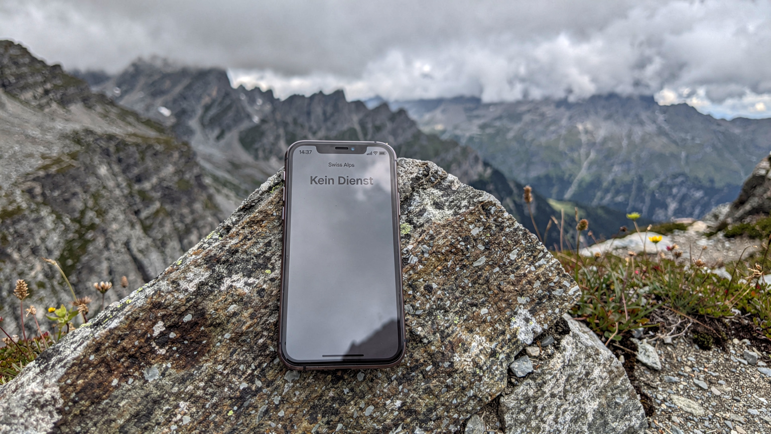 Ein Smartphone liegt auf einem zerklüfteten Felsen in den Alpen und zeigt auf dem Display 'Kein Dienst' an, was fehlende Netzabdeckung symbolisiert.
