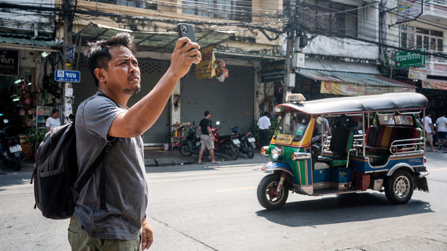 Seorang bapak sedang mencoba mencari sinyal di ponselnya, berdiri di tepi jalan yang ramai di Bangkok, dengan taksi tuk-tuk dan rambu jalan berbahasa Thailand di latar belakang.