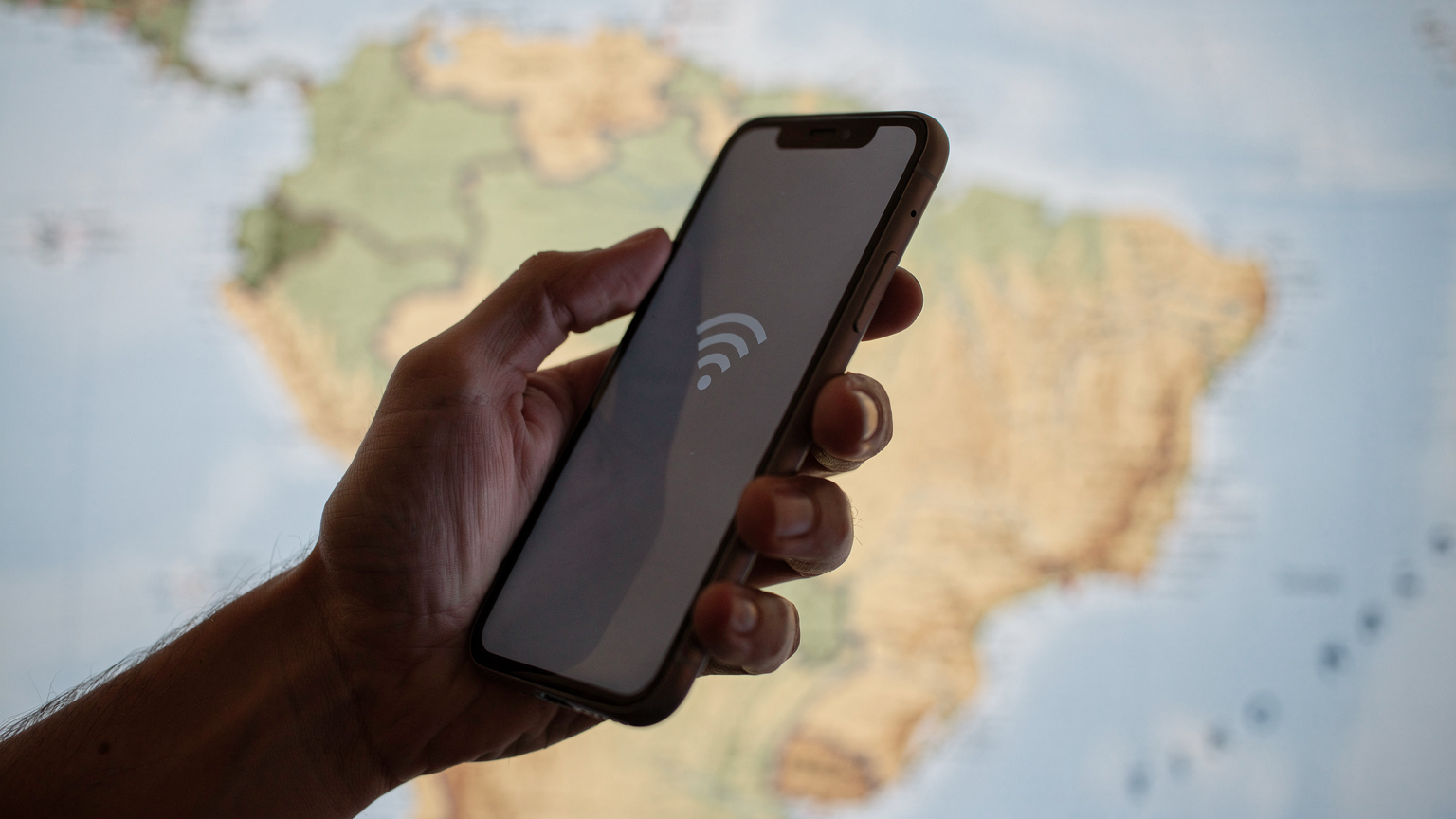 Un primo piano di una mano che tiene uno smartphone, mostrando un'icona di Wi-Fi su uno schermo leggermente illuminato, con una mappa stilizzata del Brasile in sottofondo.
