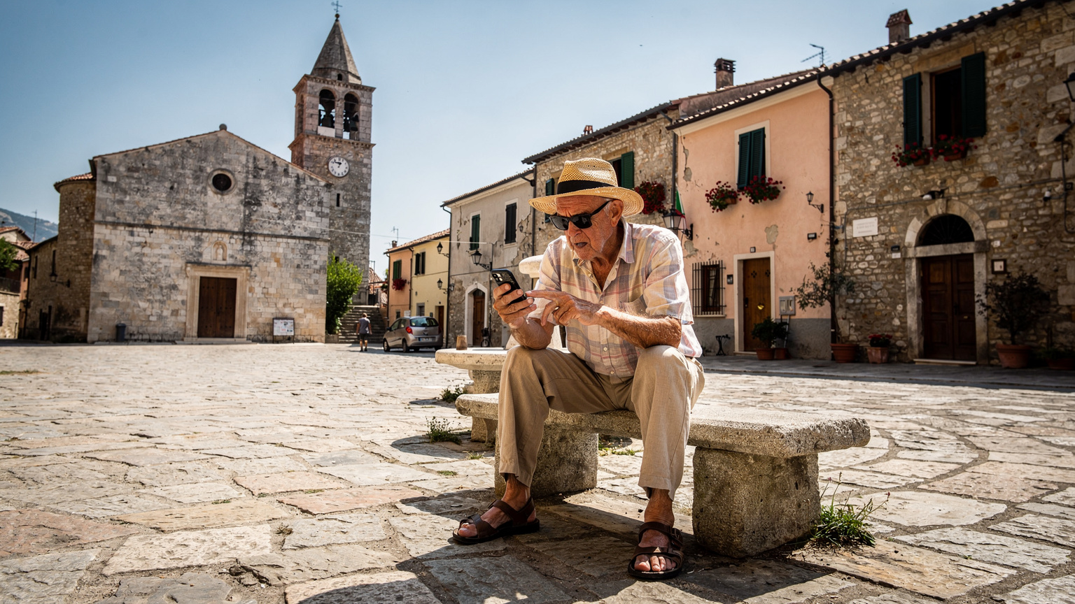Un uomo anziano, con occhiali da sole e un cappello di paglia, è seduto da solo su una panchina di pietra in una piazza assolata di un piccolo borgo italiano. Sta guardando il suo smartphone con un'espressione confusa e frustrata, tentando di trovare un segnale. Sullo sfondo, si vedono antiche case in pietra e un campanile, suggerendo un'area con infrastrutture limitate.