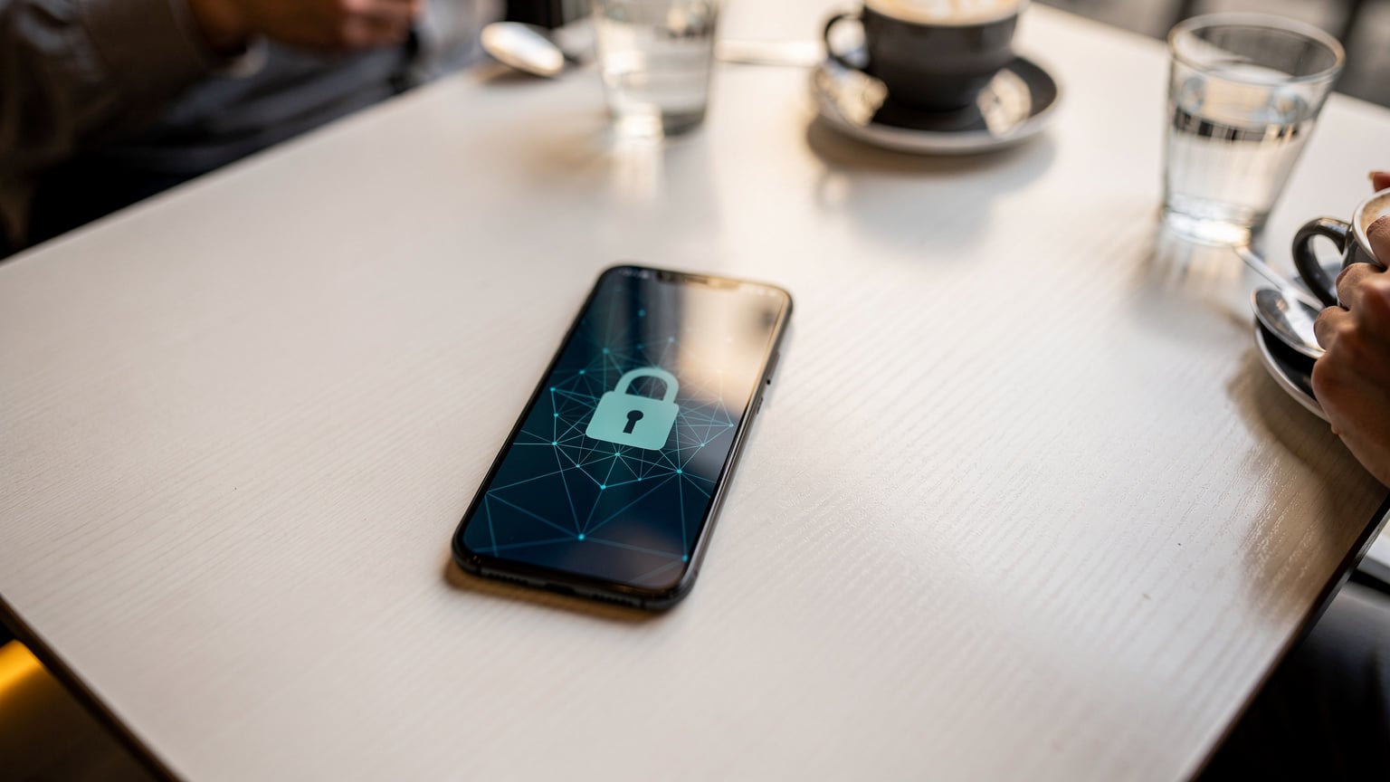 Ein Smartphone-Bildschirm zeigt ein stilisiertes Schloss-Symbol über einem abstrakten digitalen Netz, das Datensicherheit symbolisiert, während es auf einem Tisch in einem modernen Café in Pristina liegt.