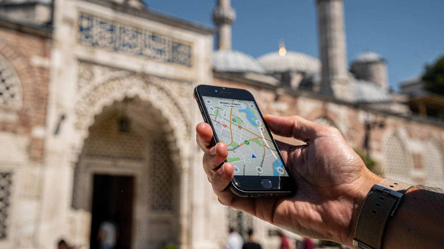 Bir gezginin eli, İstanbul'daki tarihi bir binanın önünde telefonundaki harita uygulamasını kullanıyor, ekranı hafifçe görünür, güneşli bir gün.