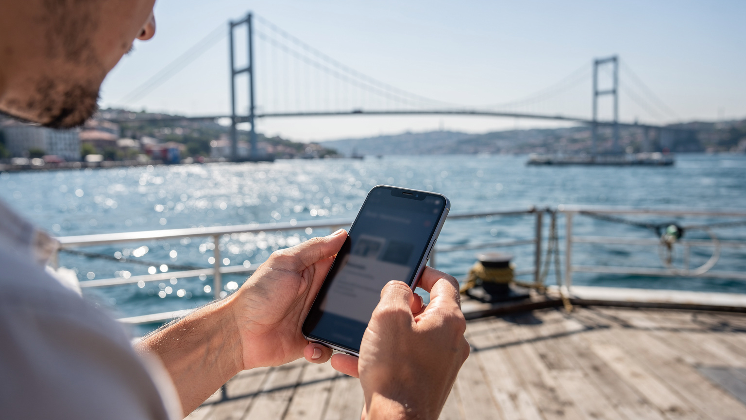 Ein Reisender hält sein Smartphone mit einer geöffneten eSIM-App in der Hand, während er auf einem Fährenterminal in Istanbul wartet, mit der Bosporus-Brücke im Hintergrund.