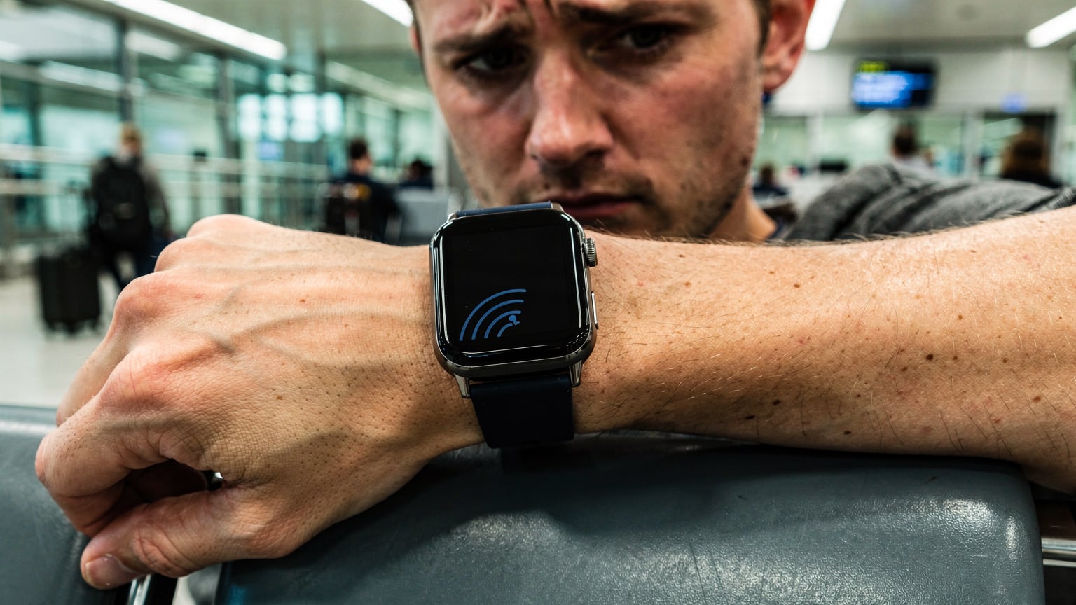 Et nærbilde av en reisende som ser bekymret på sin Apple Watch, som viser en "ingen tjeneste"-melding, mens de sitter på en flyplassbenk.