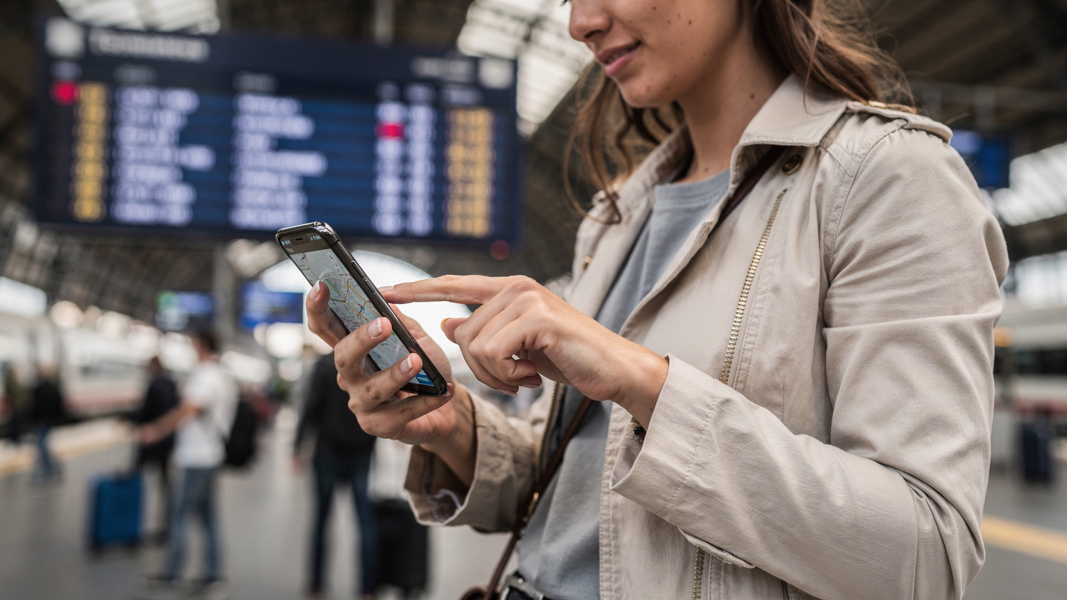 O călătoare româncă verifică o hartă digitală pe telefonul său, cu degetul pe ecran, în timp ce așteaptă un tren la Gara Termini din Roma, cu panoul electronic de sosiri/plecări în fundal estompat.