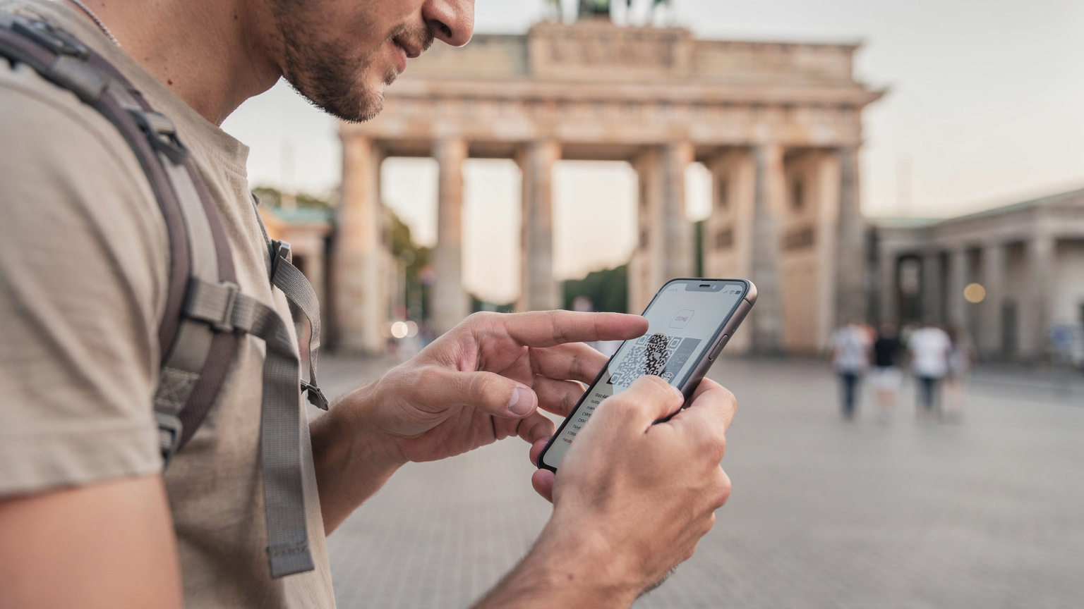 Un backpacker scanează rapid un cod QR de eSIM în fața Porții Brandenburg din Berlin, folosind lumina blândă a dimineții pentru vizibilitate, înainte de a-și continua drumul.