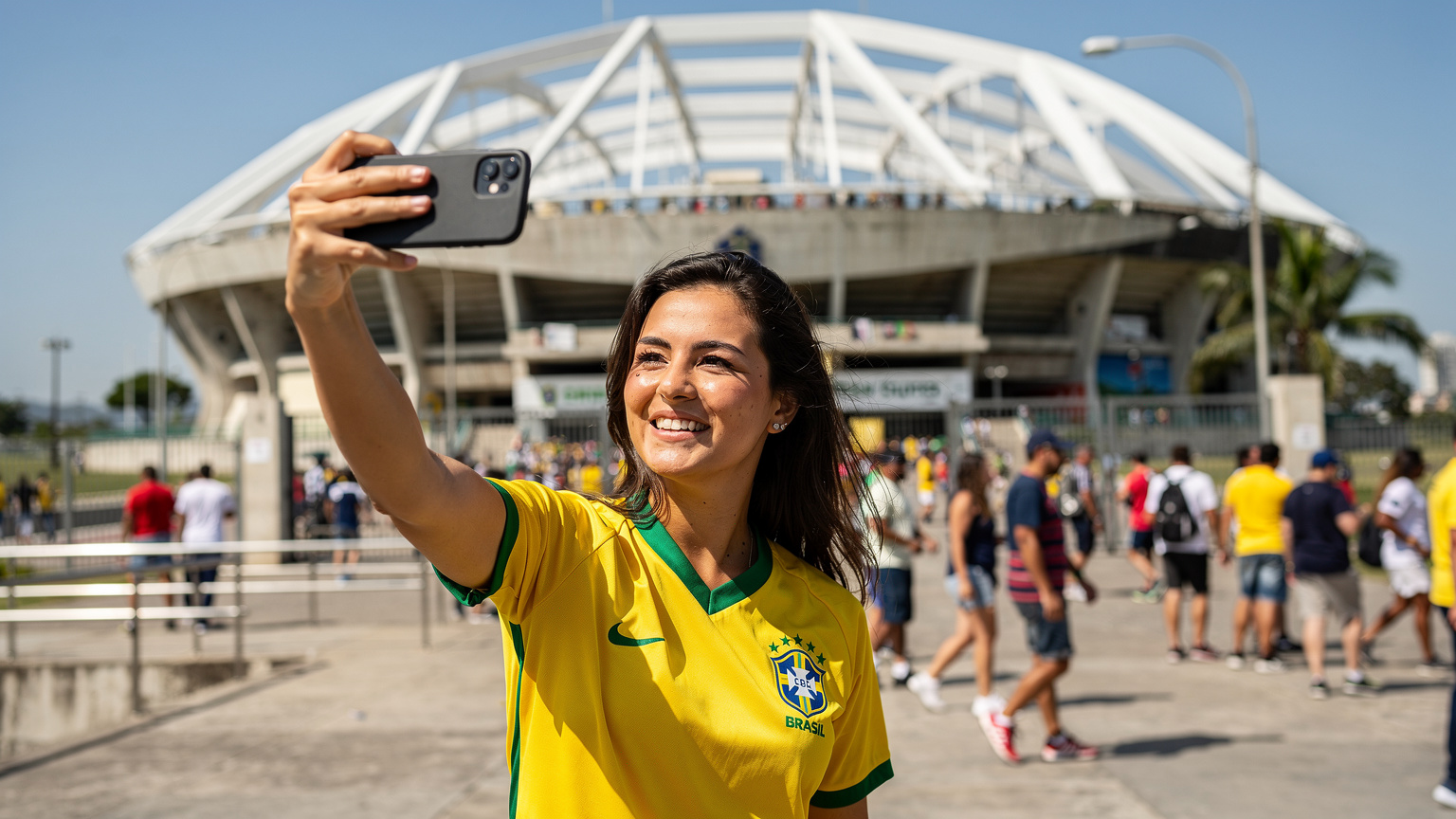 Uma torcedora sorridente, com a camisa do Brasil, usa o smartphone para tirar uma selfie na entrada do Estádio Nilton Santos, com a fachada do Engenhão ao fundo.