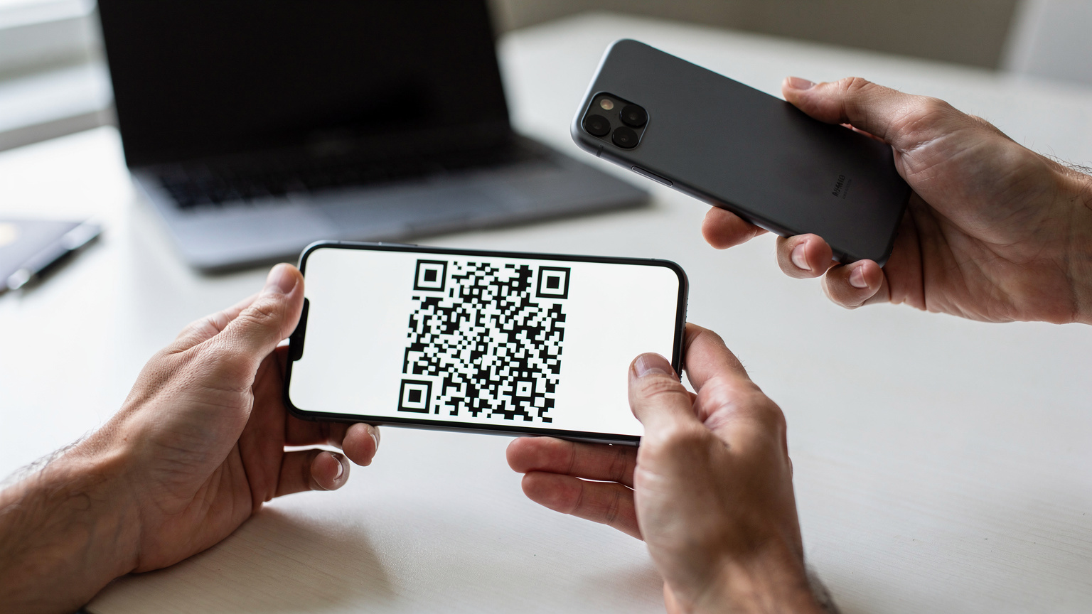 Un primo piano delle mani di una persona che tiene uno smartphone e scansiona un QR code da un altro telefono, con un laptop sullo sfondo, in un ambiente domestico o di ufficio.