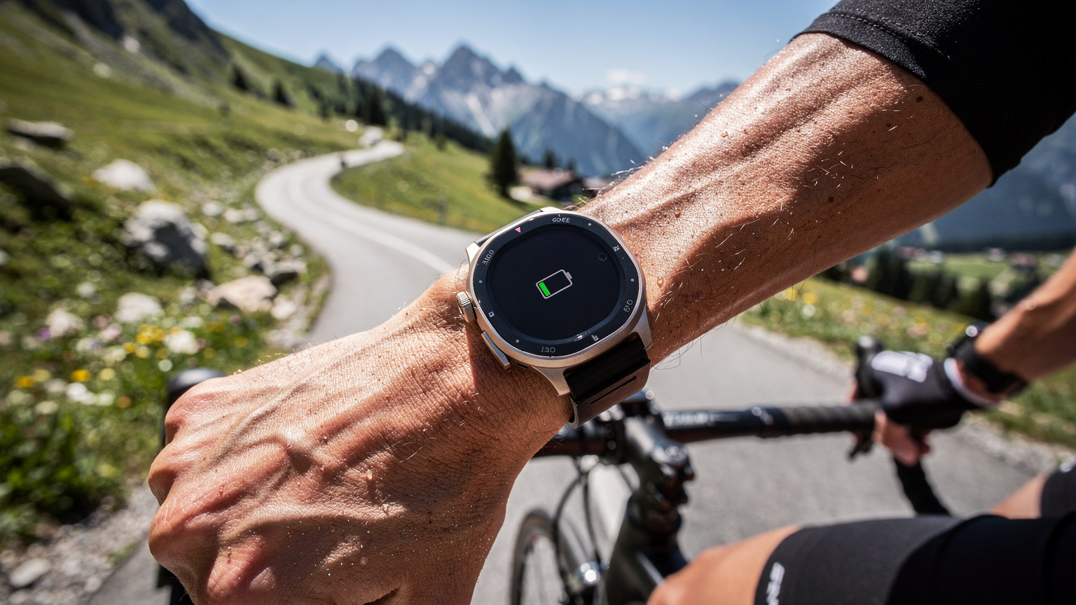 Eine detaillierte Nahaufnahme einer Apple Watch Ultra am Handgelenk eines Radfahrers, der eine Bergstraße in den bayerischen Alpen hinauffährt, mit dem Fokus auf die Uhr, die eine niedrige Akkuladung anzeigt.