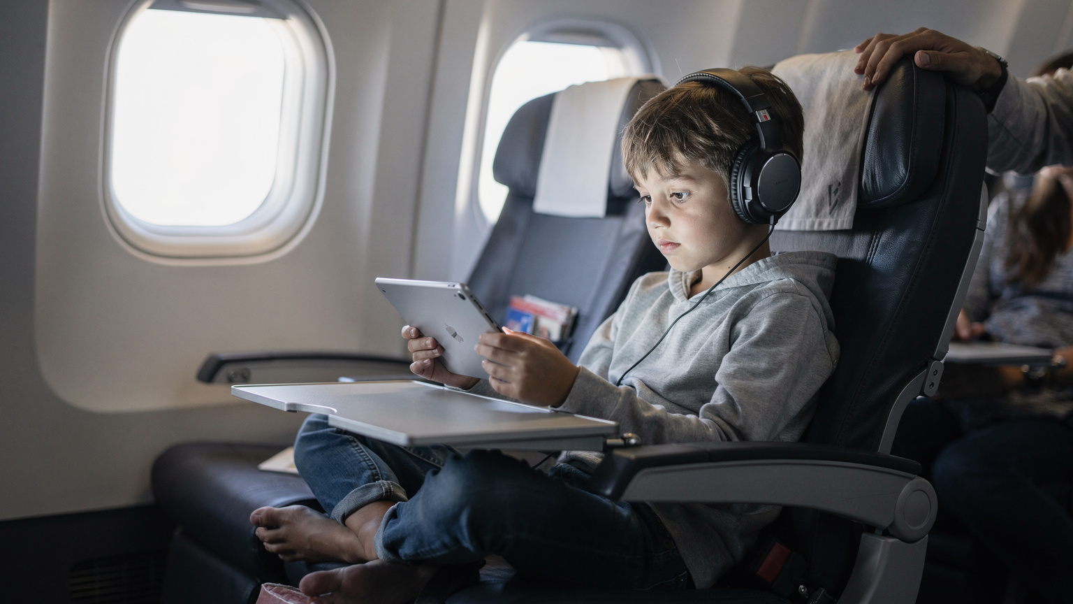 Et ungt barn sitter på et flysete og ser konsentrert på en iPad, med hodetelefoner på og en forelder som kikker over skulderen.