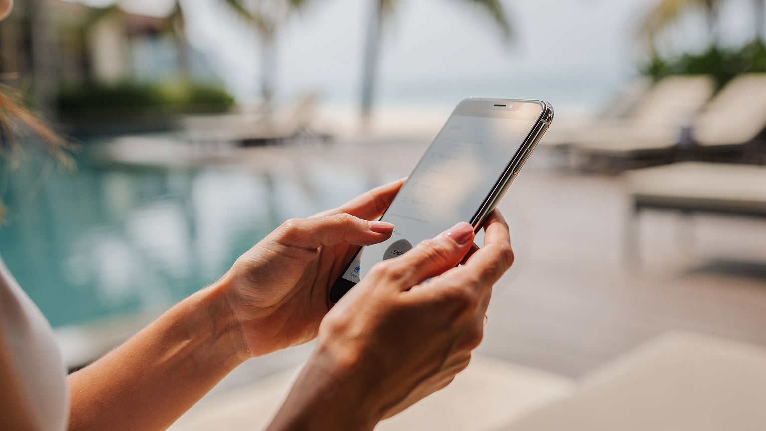 Nærbilde av en kvinnes hender som forsiktig trykker på en iPhone-skjerm, med fingrene som navigerer i innstillinger-menyen. Bakgrunnen er uskarpt, et glimt av et thailandsk feriested.