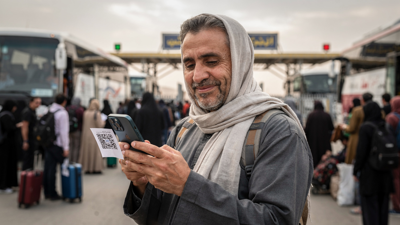یک زائر میانسال در حال اسکن کردن QR کد eSIM روی یک کاغذ در دستش, در حالی که در پس‌زمینه, انبوه جمعیت زائران و اتوبوس‌ها در گذرگاه مرزی مهران دیده می‌شود. او لبخندی بر لب دارد و گوشی‌اش در حال اتصال به شبکه است.