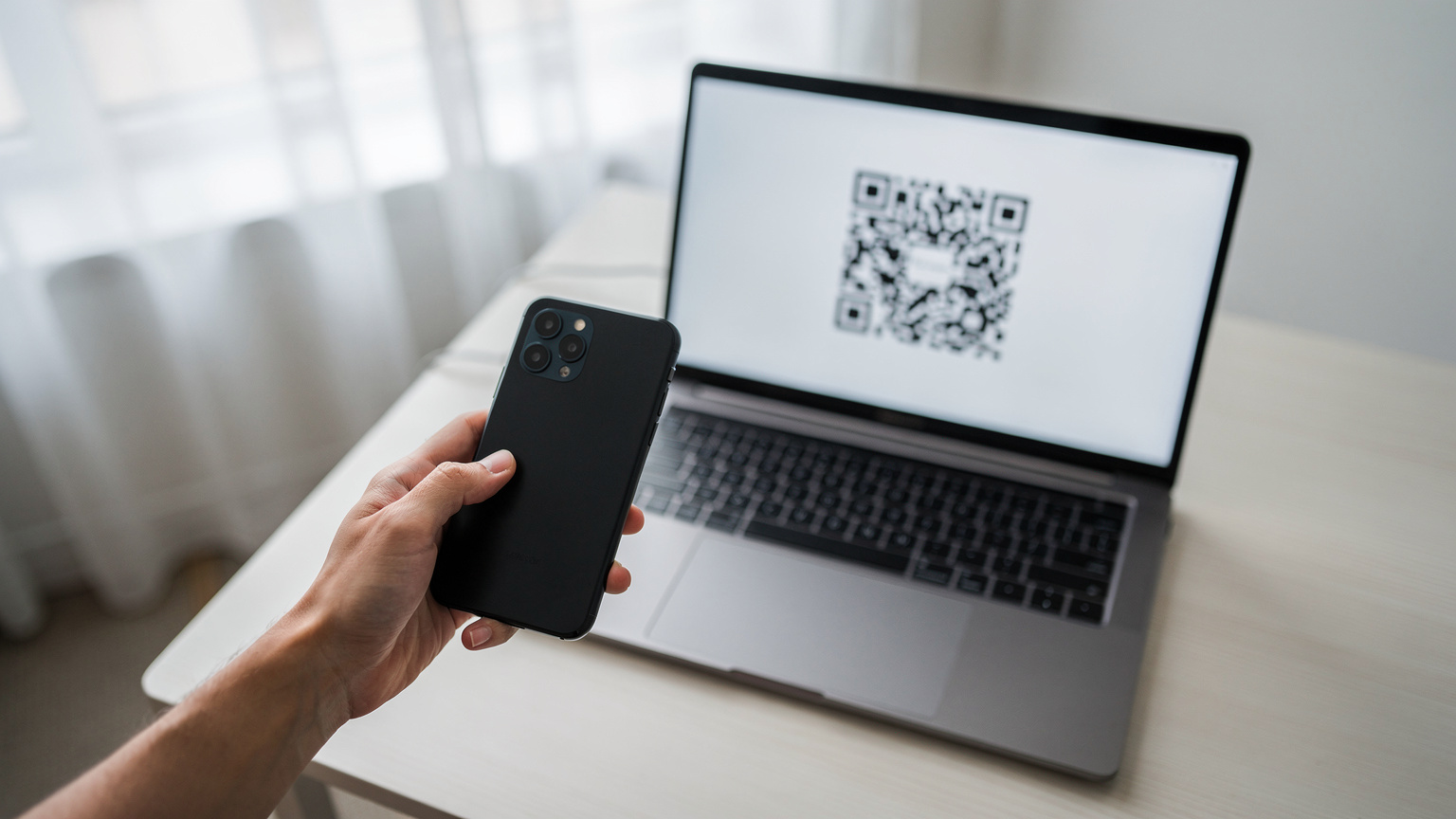Närbild på en hand som håller en smartphone och skannar en QR-kod från en bärbar datorskärm, i ett ljust, modernt rum.