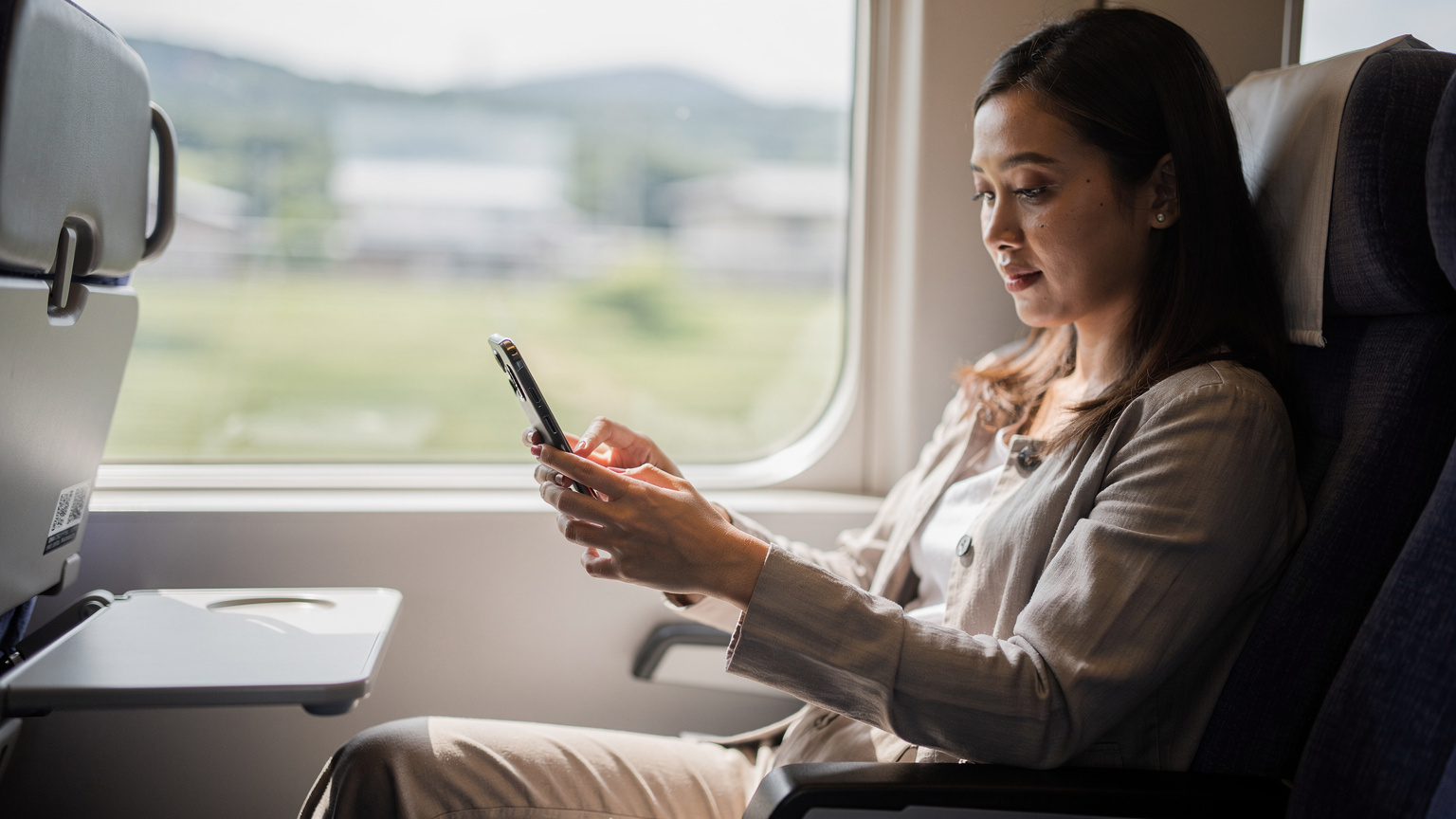 Seorang wanita muda Indonesia sedang memindai kode QR untuk mengaktifkan eSIM di ponselnya saat duduk di kursi kereta Shinkansen yang nyaman di Jepang, dengan pemandangan pedesaan yang kabur di luar jendela.