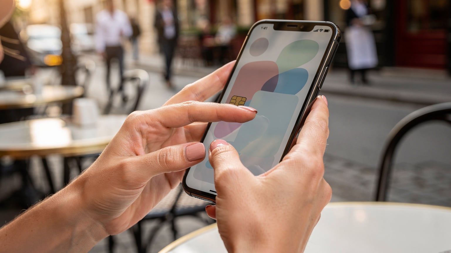 Nahaufnahme der Hände einer jungen Frau, die auf einem Smartphone-Bildschirm eine eSIM-App bedient, im unscharfen Hintergrund ein typisches Pariser Bistro mit Passanten.