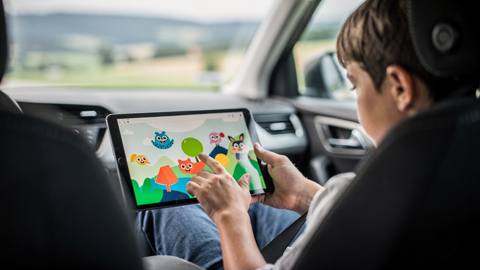 Zblízka záběr na ruce dospělého a dítěte, jak společně drží tablet, na kterém je spuštěná offline hra, zatímco sedí v autě během dlouhé cesty.