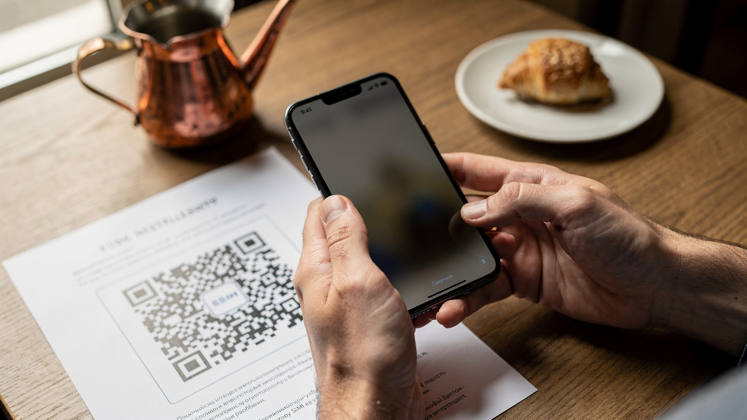 Κοντινό πλάνο ενός χεριού που κρατά ένα smartphone με την οθόνη να δείχνει τη διαδικασία σάρωσης ενός κωδικού QR για την εγκατάσταση μιας eSIM, με ένα φλιτζάνι καφέ στο παρασκήνιο.
