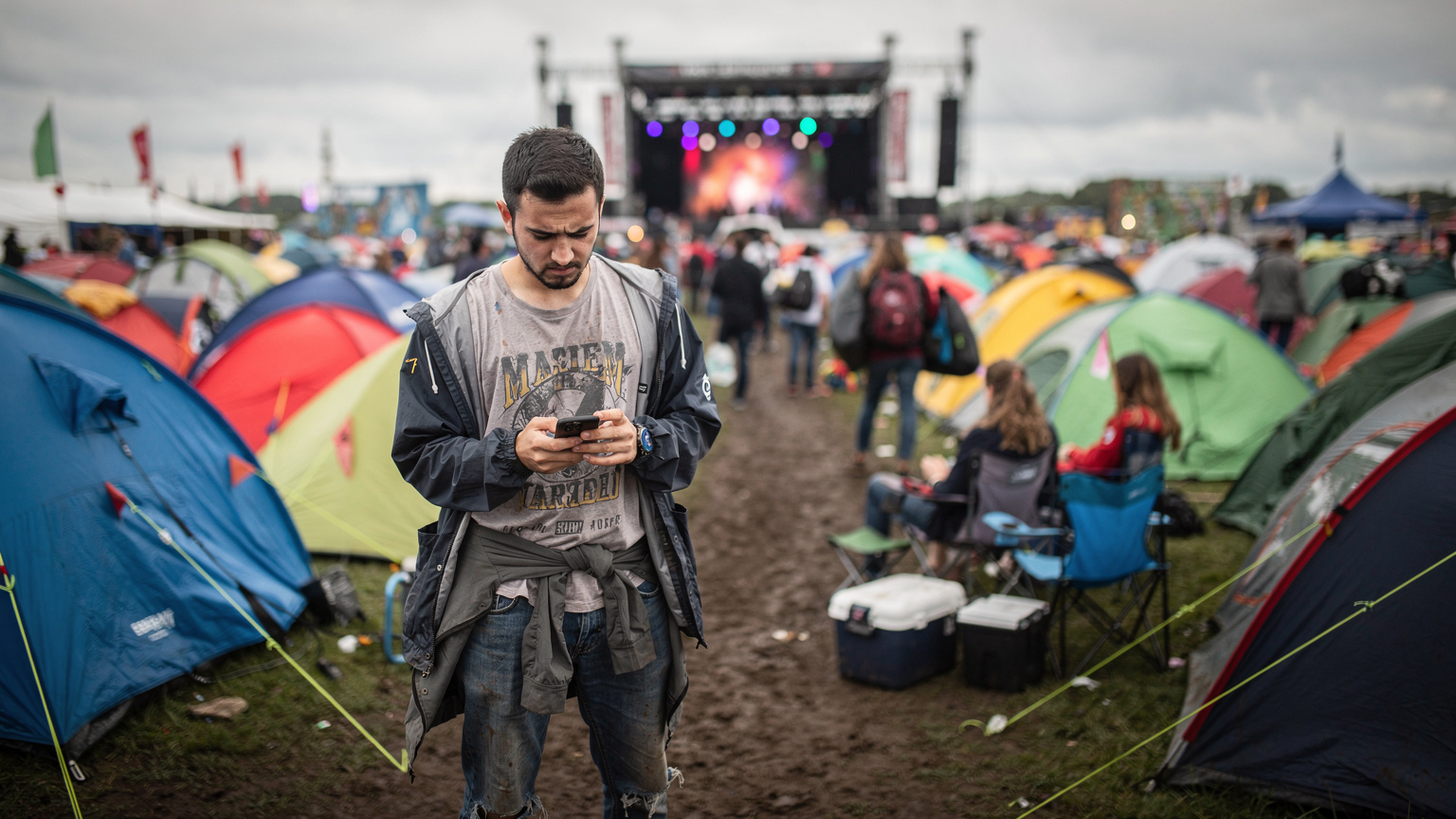 Glastonbury Festivali'nde bir Türk genci, çadırların arasında arkadaşlarıyla buluşmak için telefonundan haritaya bakarken, arka planda devasa müzik sahnelerinden biri ve coşkulu kalabalık görülüyor.