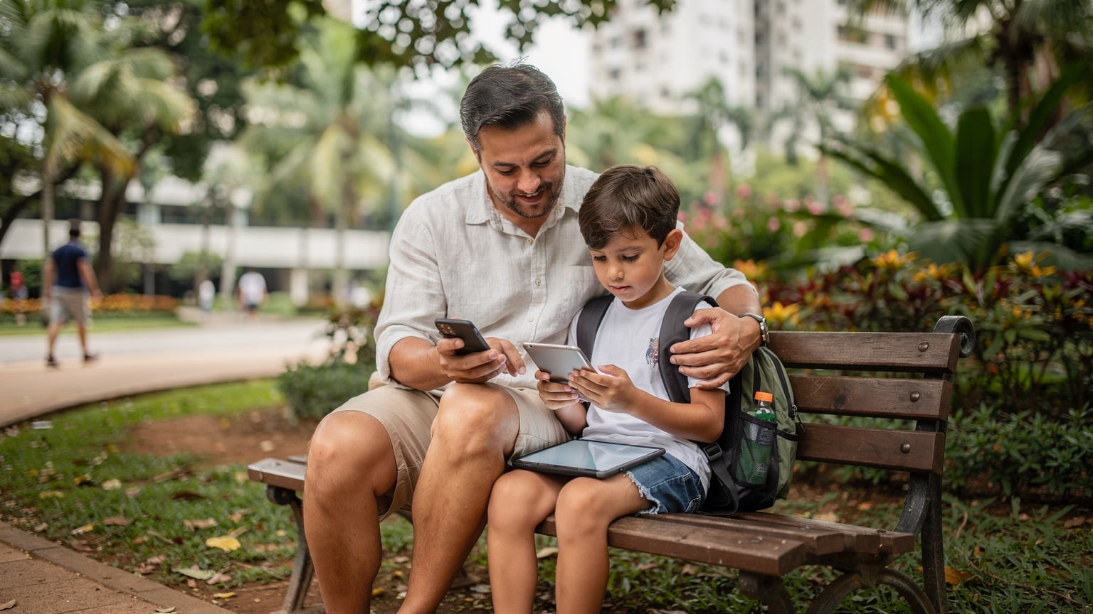 Un parent est assis sur un banc dans un parc urbain brésilien, tenant son smartphone et aidant son enfant à installer un jeu sur une tablette. Les deux sont concentrés sur les appareils, avec une ambiance détendue et verdoyante en arrière-plan.