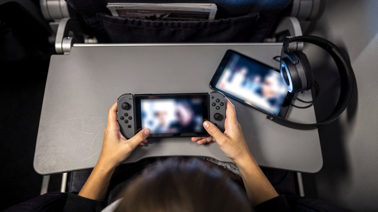 Gros plan sur les mains d'un enfant de 10 ans jouant à un jeu sur une Nintendo Switch lors d'un long vol. À côté de la console, on voit une tablette affichant un film et un casque audio. L'ambiance est calme et concentrée.
