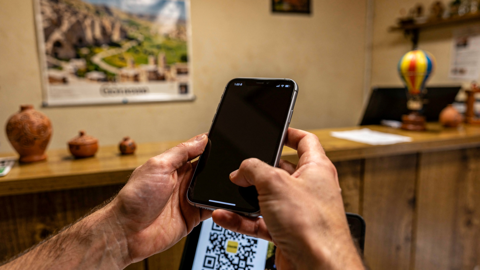 Un primer plano de las manos de un viajero utilizando su smartphone para escanear un código QR de eSIM, con un fondo ligeramente desenfocado de un mostrador de recepción de hostel en Capadocia.