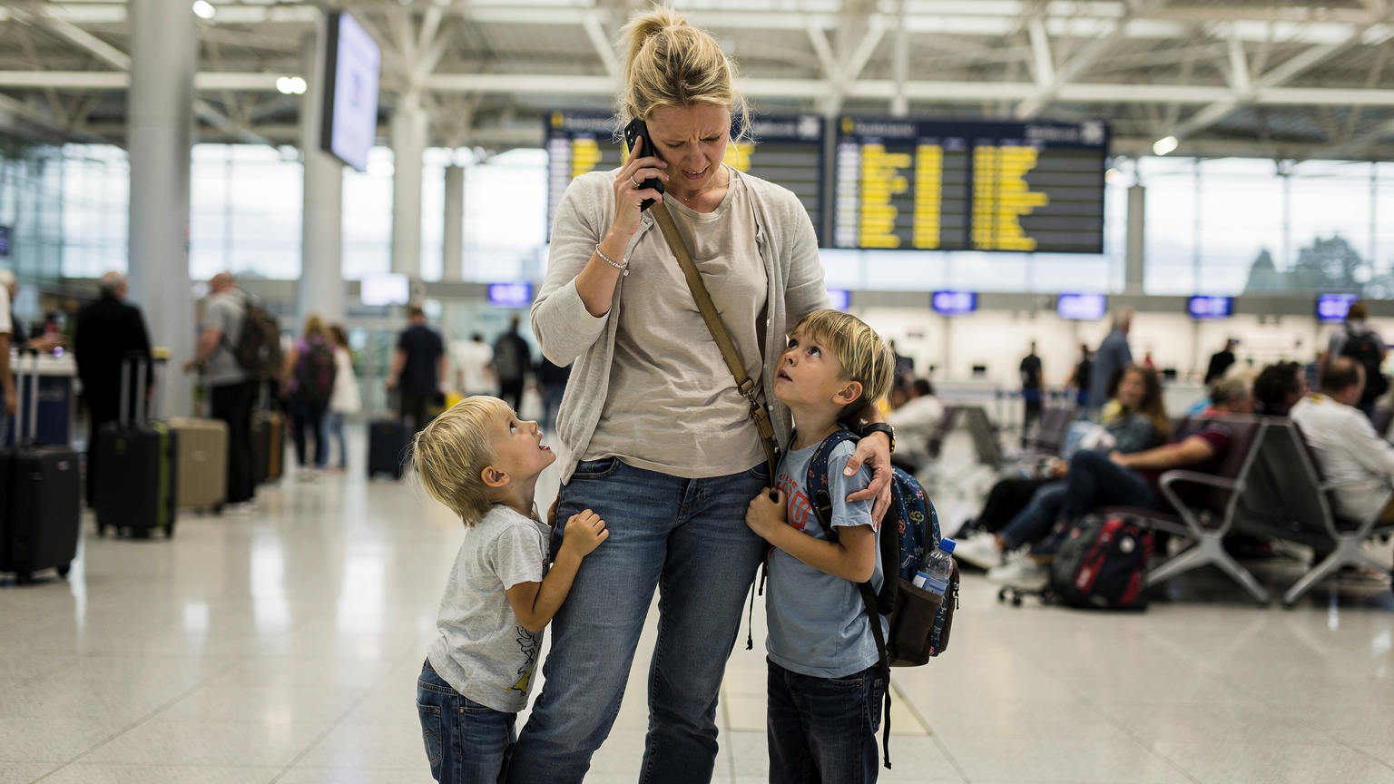 En stressad mamma med två barn som drar i henne, där hon försöker få kontakt med kundtjänst på sin telefon på en livlig flygplats.