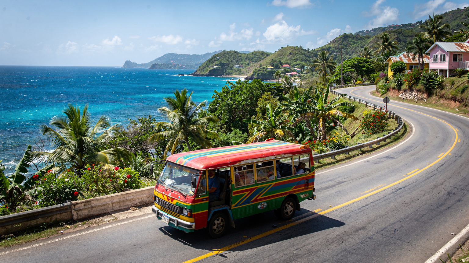 A mini-bus, characteristic of Grenada's public transport, navigating a winding coastal road with lush green hills in the background.