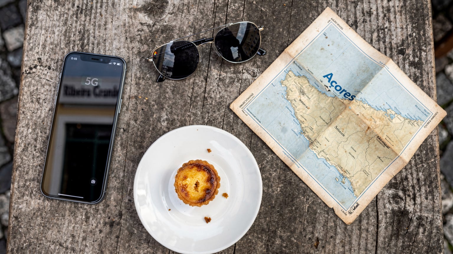 Uma vista aérea de uma mesa de café na Ribeira Grande, Açores, com um mapa, um smartphone com eSIM ativo, óculos de sol e um bolo de queijo local.