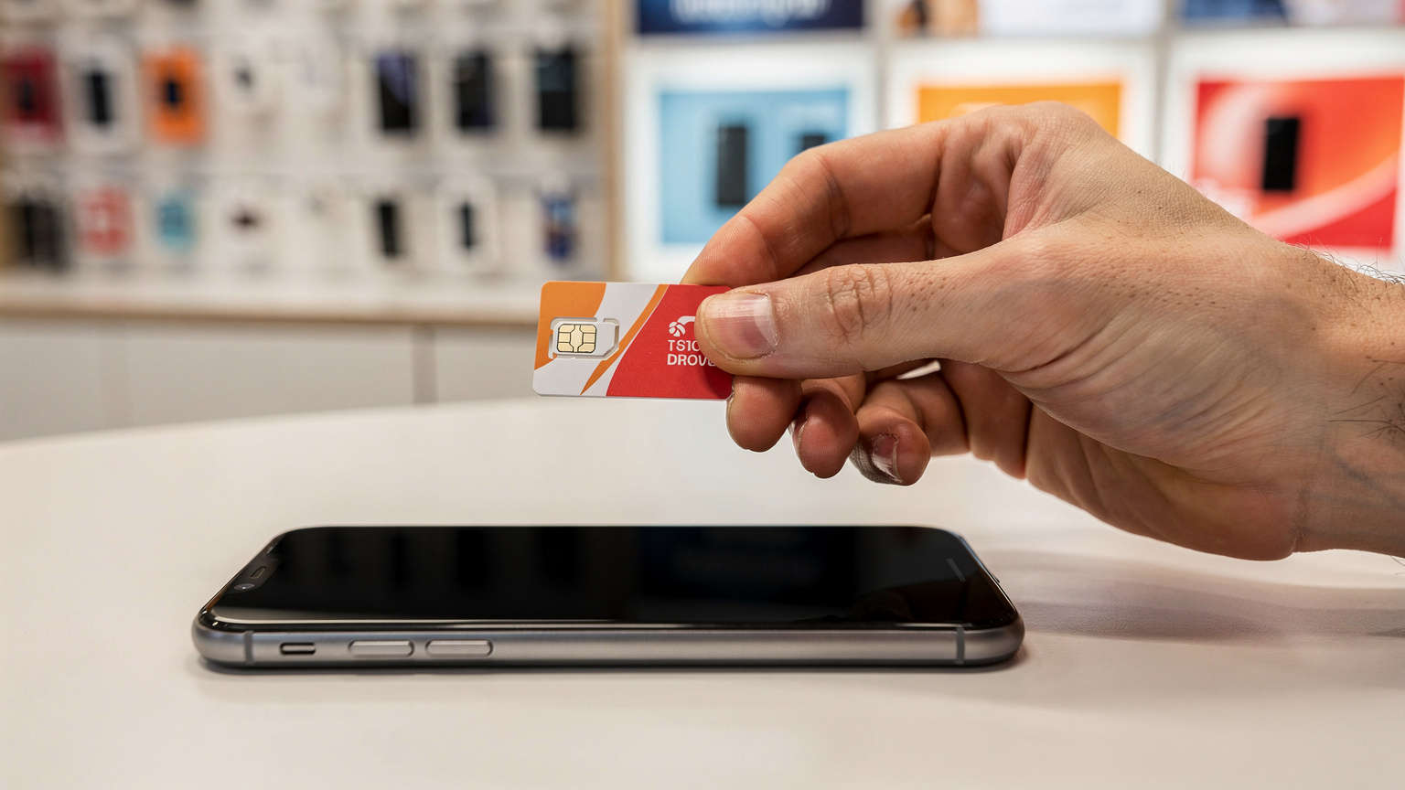 Une main tient une carte SIM physique d'un opérateur mobile français, avec un smartphone posé sur une table à côté, dans une boutique de téléphonie mobile à Paris, suggérant le dilemme du choix.
