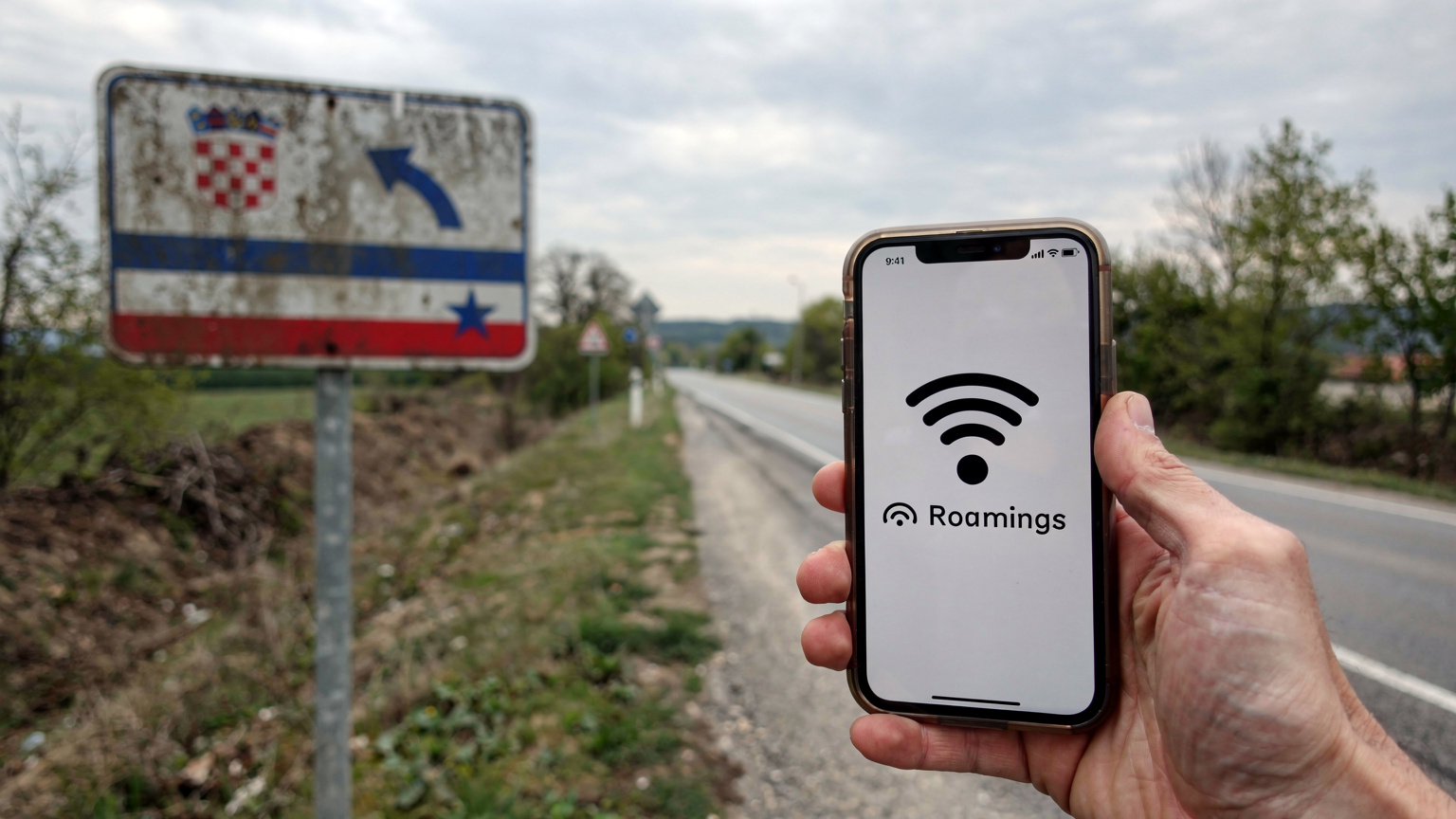 Un primo piano di un cartello stradale che indica i confini tra Croazia e Bosnia-Erzegovina, con una mano che tiene uno smartphone in primo piano, mostrando un'icona di roaming attivo.