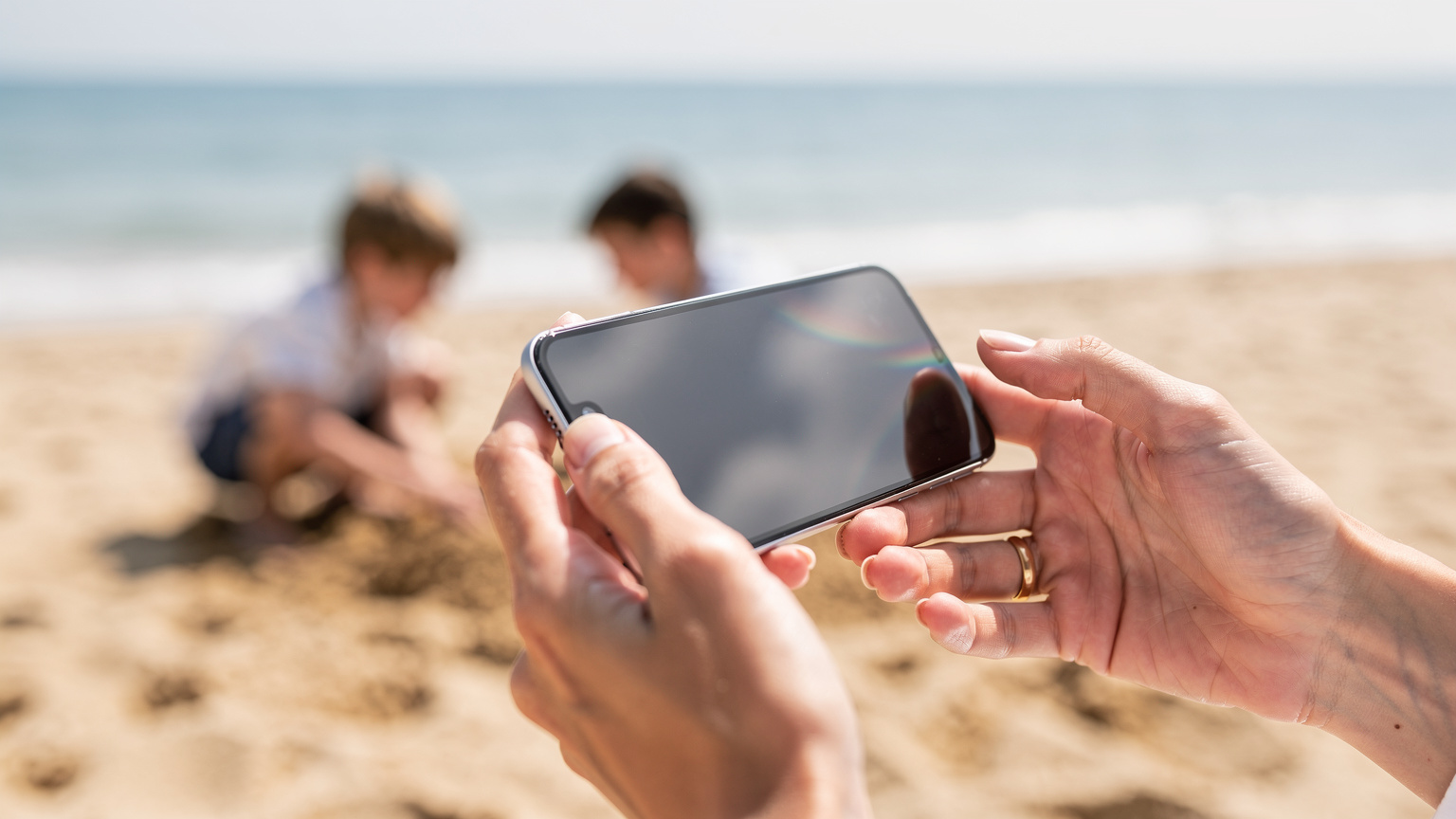 Крупний план рук мами, яка налаштовує eSIM на смартфоні, на задньому плані видно дітей, які граються на пляжі.