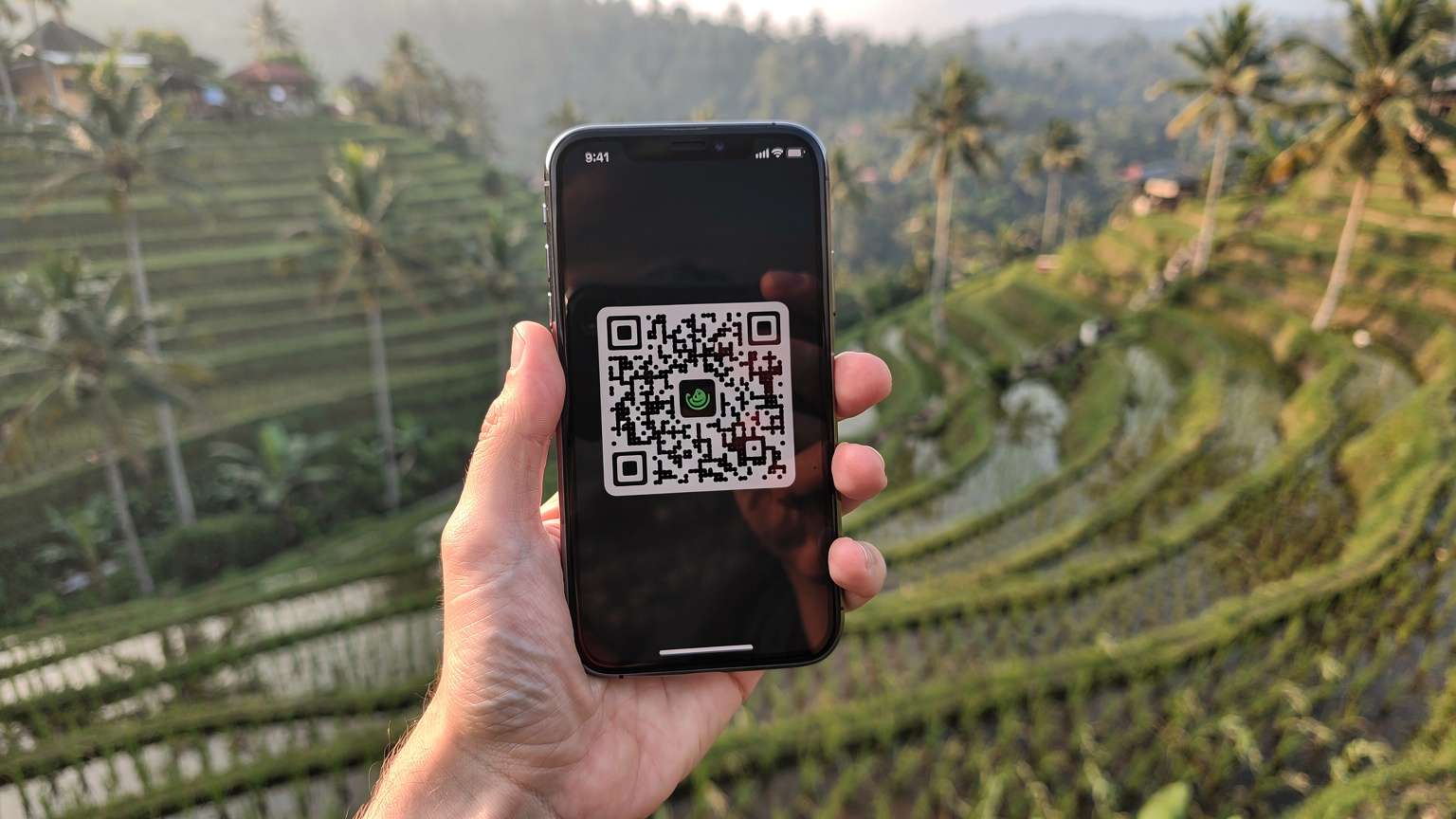 Een hand houdt een smartphone vast die een QR-code op het scherm toont, met de weelderige groene rijstvelden van Jatiluwih op Bali op de achtergrond, wazig maar herkenbaar.