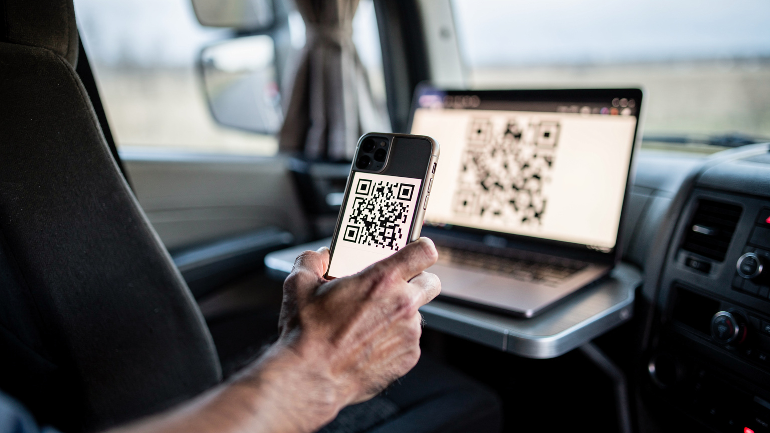 Ръка, сканираща QR код на екрана на лаптоп с камера на смартфон, в кабината на камион, паркиран на почивка.