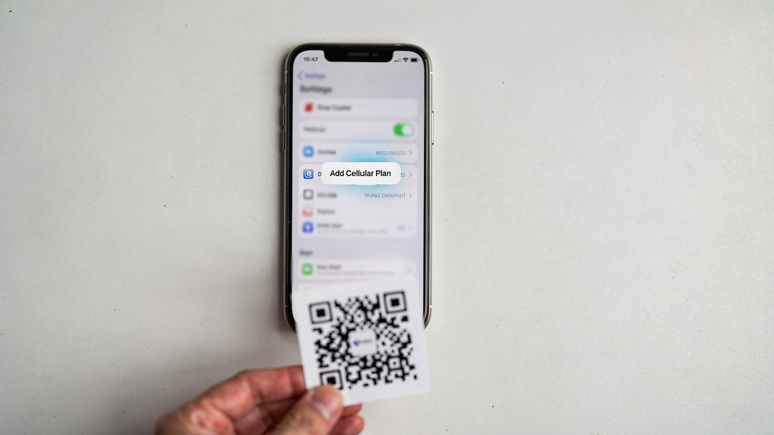 Um smartphone exibindo a tela de configurações de rede celular, com a opção de "Adicionar Plano Celular" destacada, enquanto uma pessoa escaneia um QR code impresso.