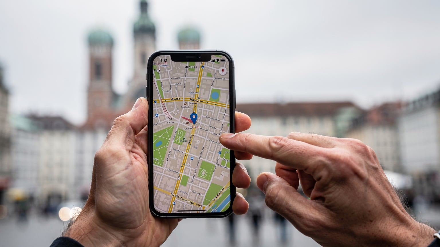 Eine Nahaufnahme zeigt die Hände einer Person, die auf ihrem Smartphone eine Karte von München betrachtet, während im Hintergrund verschwommen die Frauenkirche zu sehen ist.