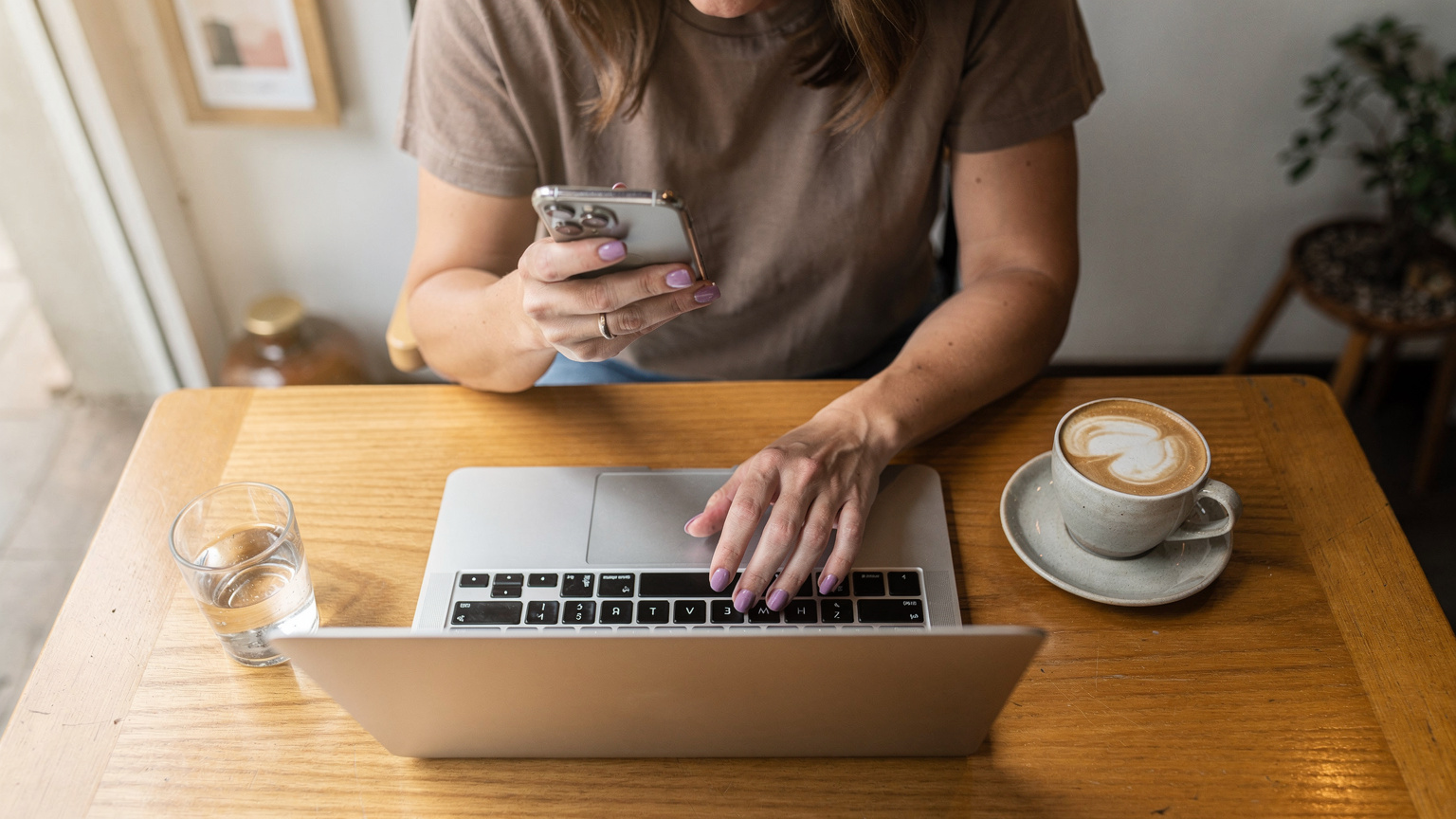 Una mujer sentada en una moderna cafetería con su laptop y café, revisa su teléfono comparando diferentes planes de eSIM en la pantalla, con un gesto pensativo.