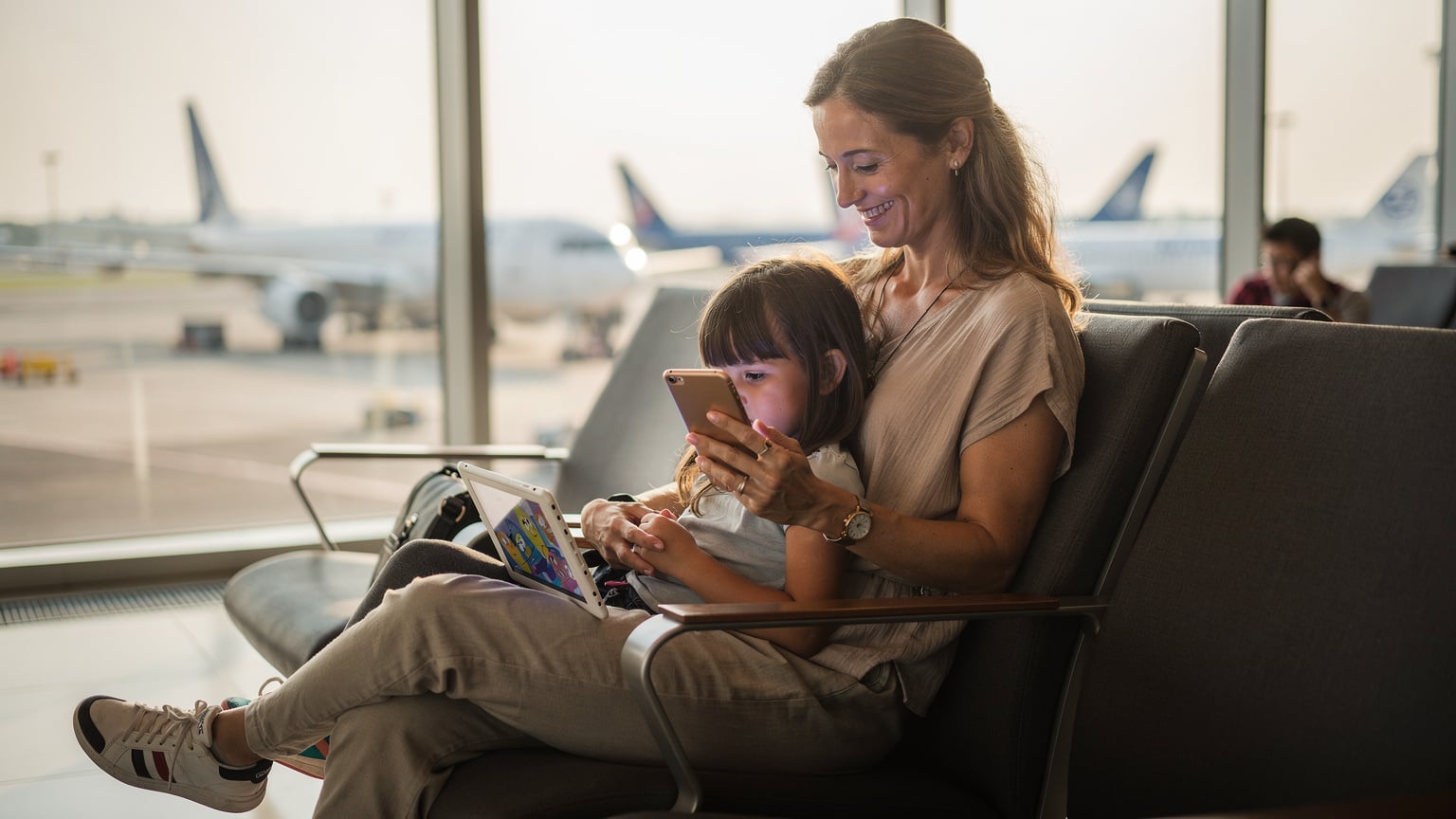 Seorang ibu tersenyum lega sambil memegang iPhone di tangan kirinya dan menggandeng tangan anak perempuannya yang sedang asyik menonton kartun di iPad di sebuah lounge keberangkatan Bandara Internasional Soekarno-Hatta yang modern dan terang.