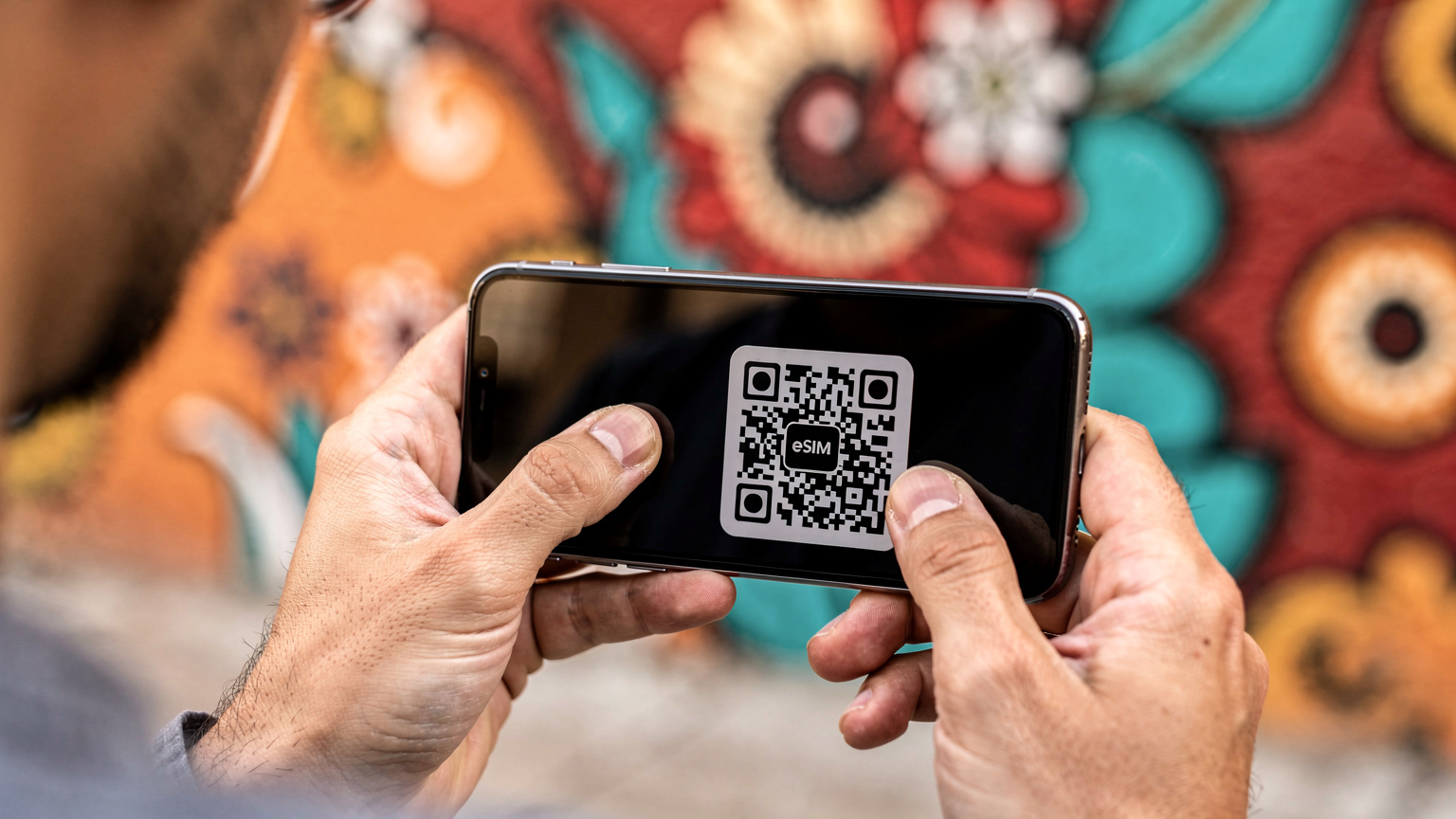 A close-up shot of hands using a smartphone to scan a QR code for eSIM activation, with a blurred background of a vibrant Mexican street art mural in Oaxaca.