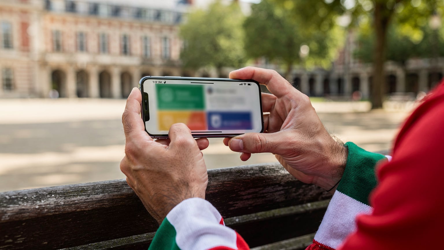 Un primo piano delle mani di un tifoso italiano che confronta i prezzi dei piani dati eSIM sul suo smartphone, con diverse schede aperte che mostrano offerte competitive, mentre è seduto su una panchina a Place des Vosges.