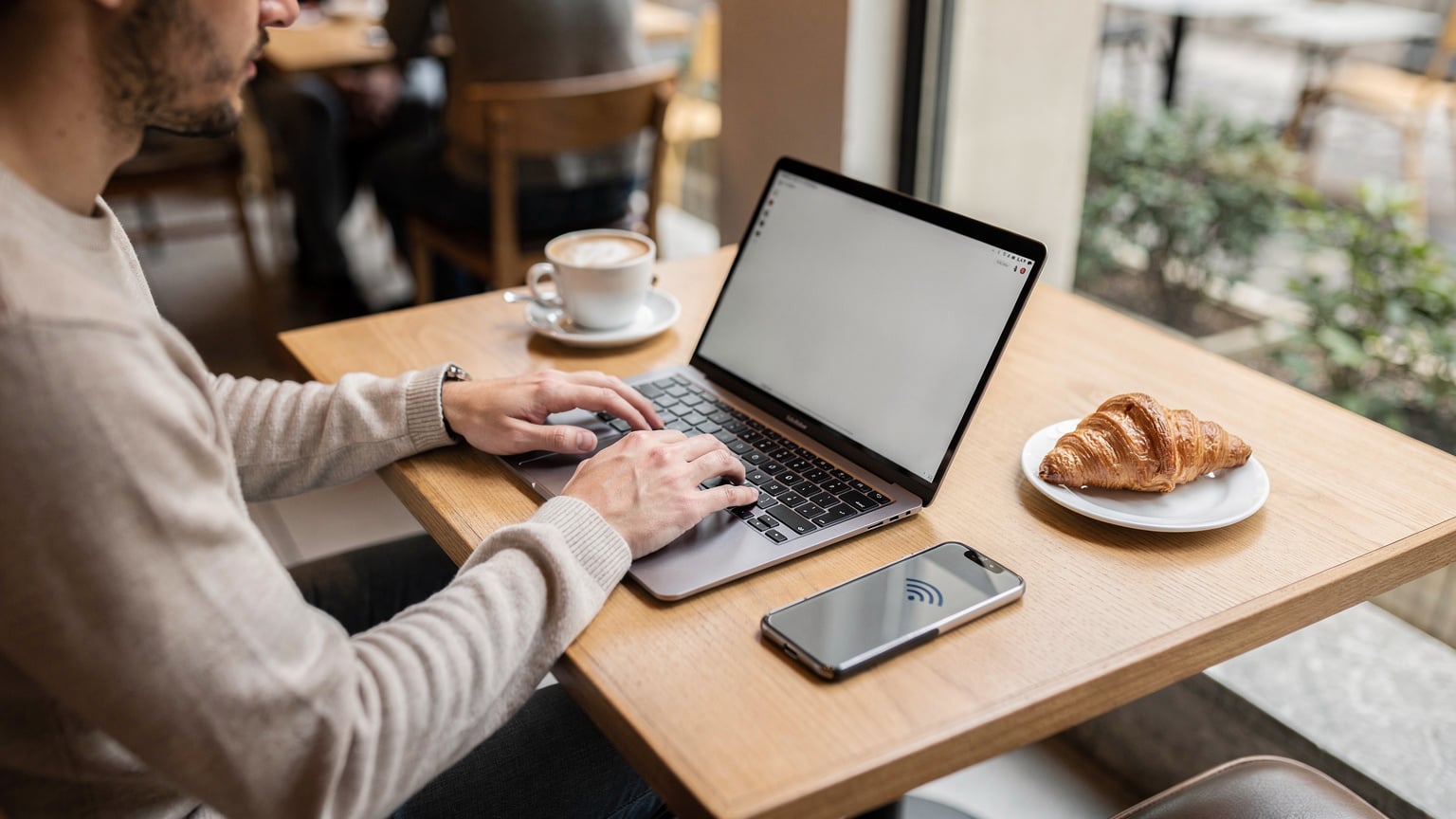 Un nomade digitale con un laptop aperto e il suo smartphone, che mostra un'icona Wi-Fi, sta lavorando tranquillamente seduto a un tavolo del Café Kitsuné, con un caffè e un croissant.