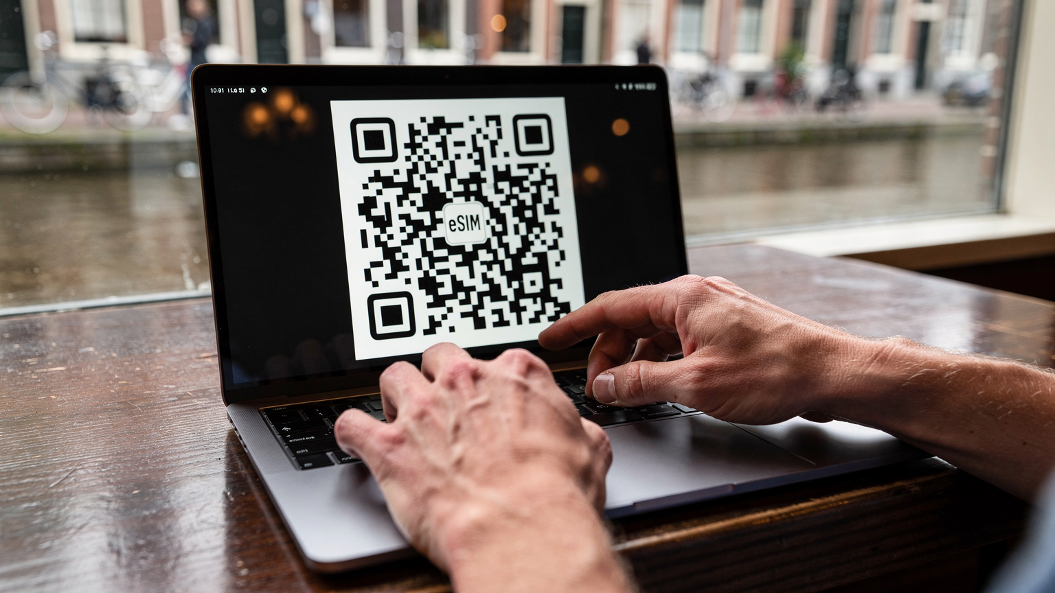 A close-up of a pair of hands, likely a parent's, gently tapping on a laptop screen with an eSIM activation QR code visible, set against the blurred background of a cozy cafe in Amsterdam's Jordaan district, with a canal visible outside the window.