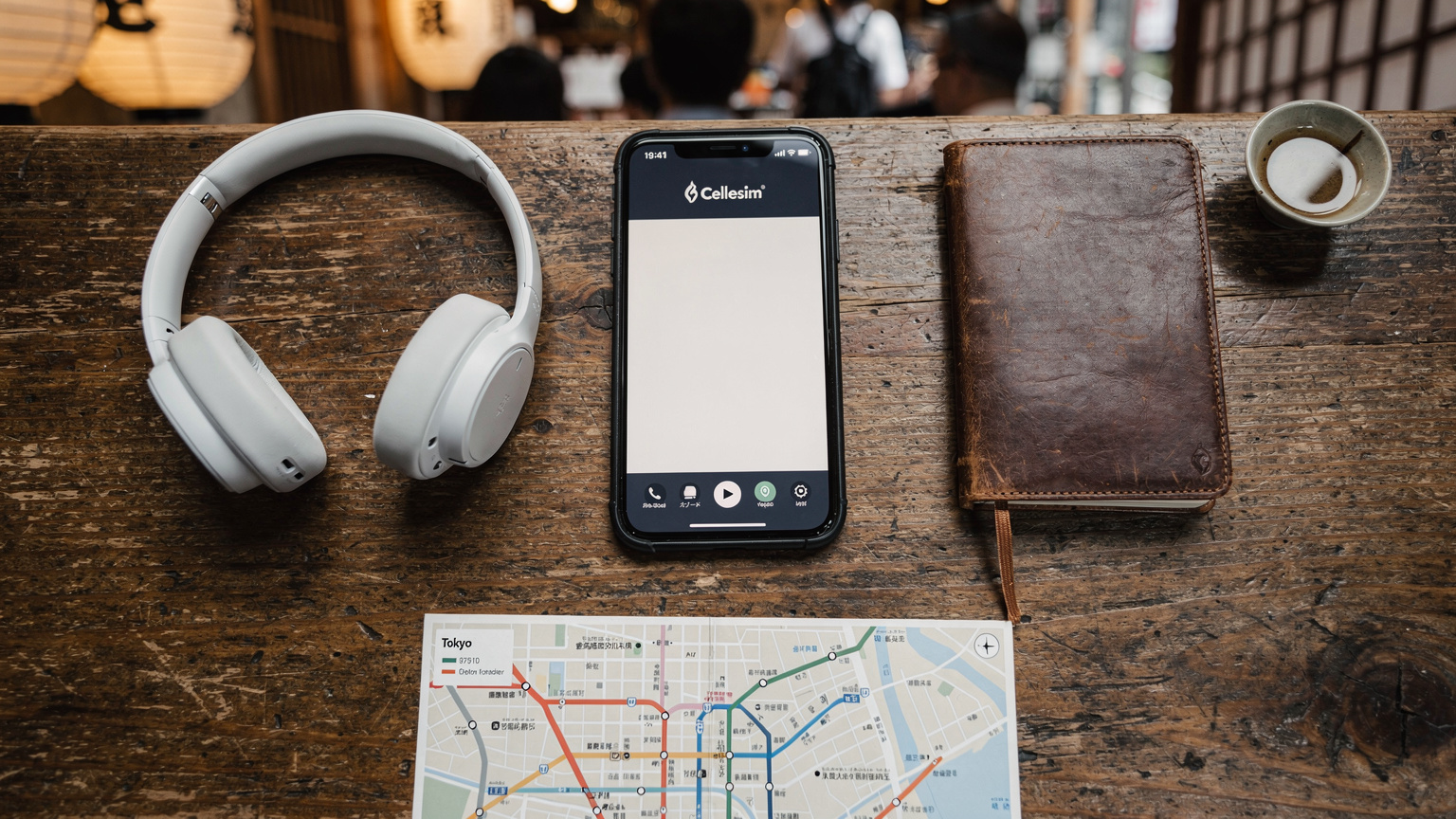 An overhead flat-lay of travel essentials: a sleek smartphone displaying a Cellesim app, a passport, a pair of wireless headphones, a small travel journal, and a local transit map, all neatly arranged on a rustic wooden table in a bustling cafe in Shibuya, Tokyo.