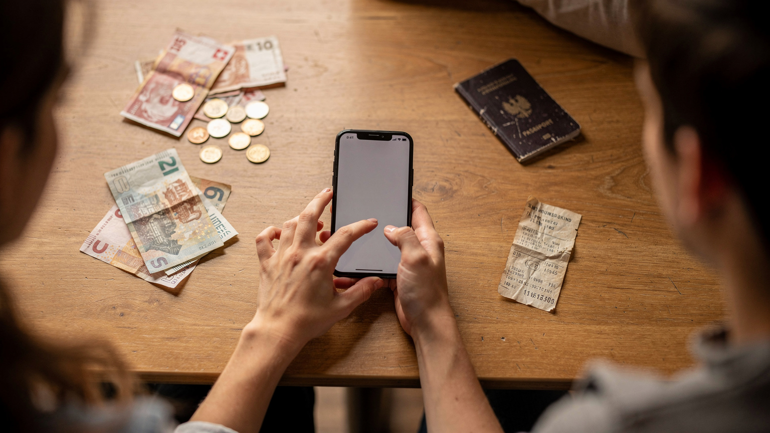 A female traveler's hands holding a smartphone, comparing various Cellesim eSIM plans for Switzerland, with a scatter of Swiss Franc notes and coins on a hostel common room table, emphasizing budgeting.