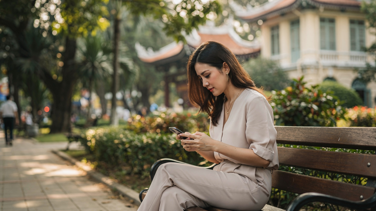 Một phụ nữ trẻ đang ngồi trên ghế đá tại công viên Tao Đàn, Thành phố Hồ Chí Minh, chăm chú nhìn vào chiếc iPhone của mình để kiểm tra cài đặt eSIM.
