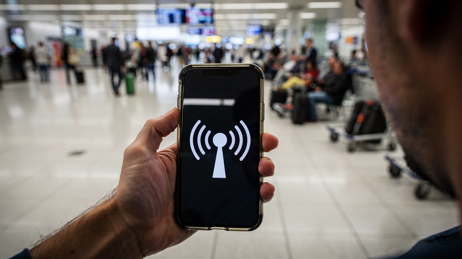 Ręka trzymająca smartfon z wyraźnym powiadomieniem o braku zasięgu, z tłem zatłoczonego lotniska i zaniepokojoną miną podróżnego.