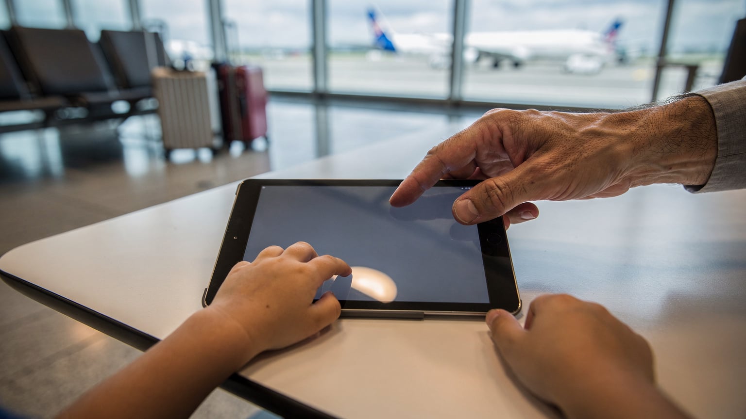 Ένα παιδικό χέρι αγγίζει ένα tablet, ενώ ένα χέρι ενήλικα κατευθύνει την οθόνη, κατεβάζοντας μια ταινία για offline προβολή στο αεροδρόμιο Charles de Gaulle.