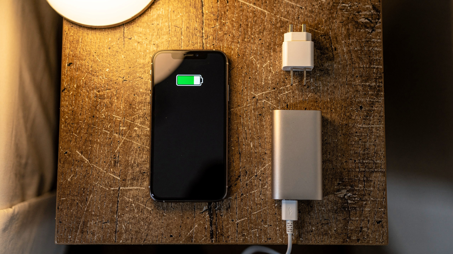 Un smartphone affichant une notification de faible batterie, posé sur une petite table en bois avec un adaptateur de prise universel et un chargeur portable à côté, dans une chambre d'hôtel modeste à Pattaya.