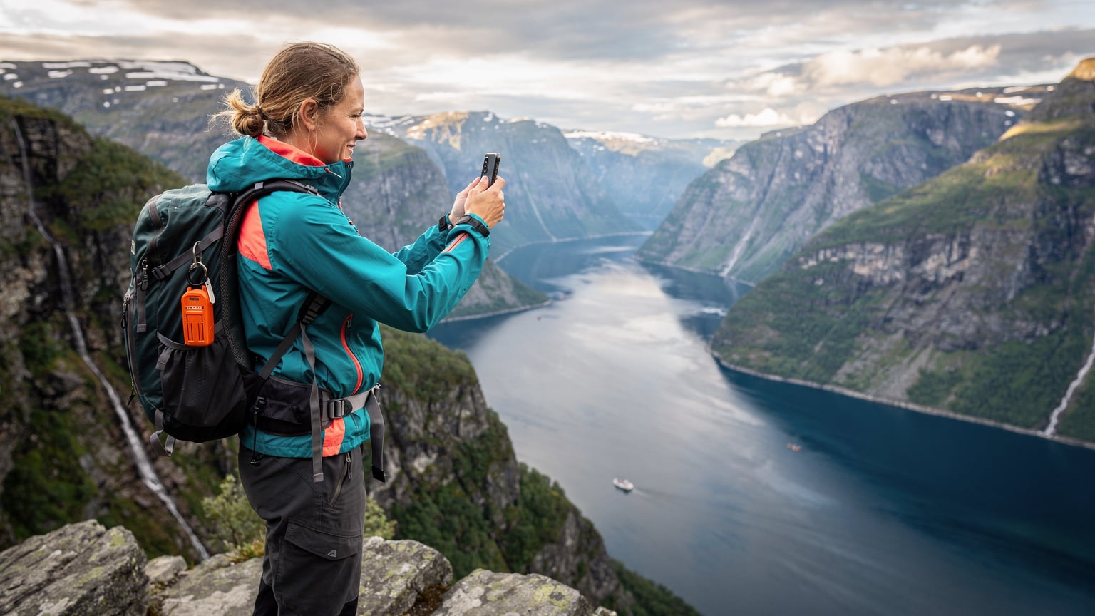 En kvinne med turbukse og vindjakke tar et bilde av en vakker fjordutsikt med sin smarttelefon, mens hun har et nødpeilesender på ryggsekken.