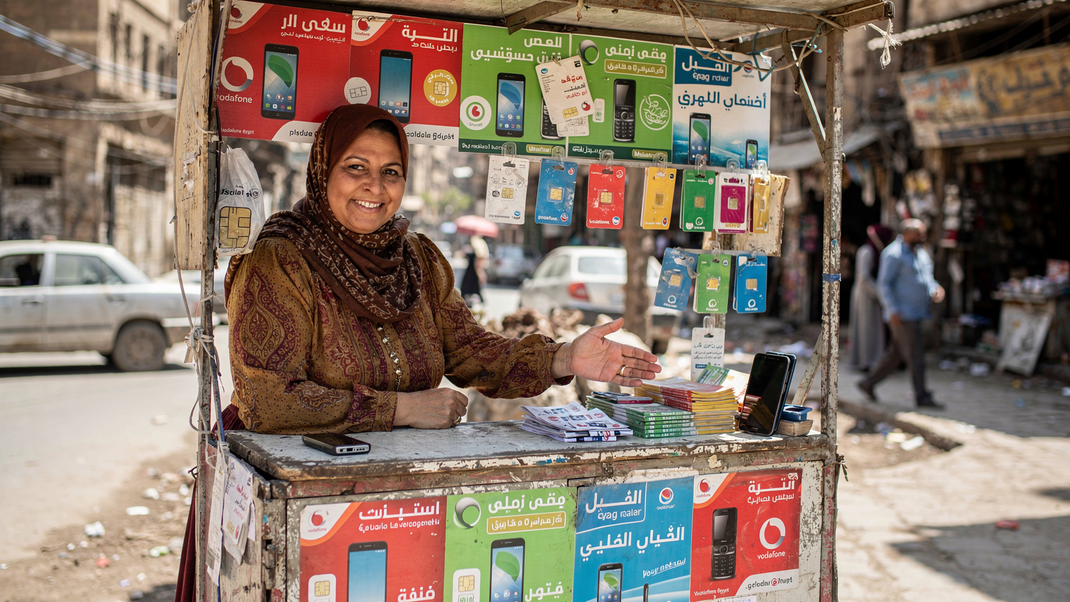 Une vendeuse souriante dans un petit kiosque de rue vendant des cartes SIM et des recharges téléphoniques au Caire, avec des panneaux publicitaires colorés en arabe.