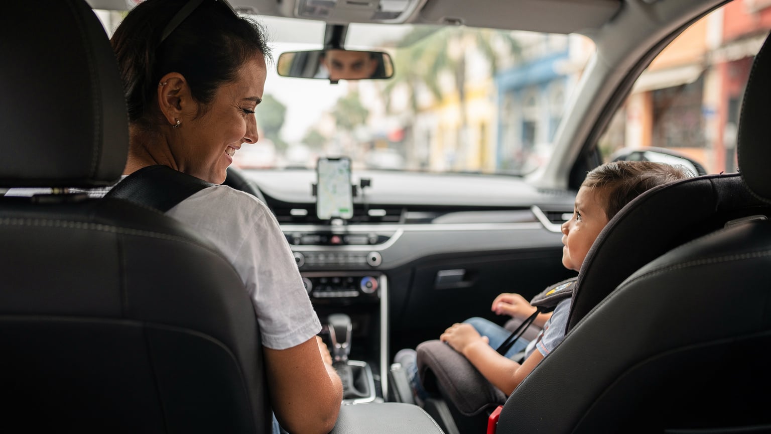 Родитель с ребенком, сидящим в детском кресле, в салоне Uber, смартфон родителя с активной навигацией на приборной панели.